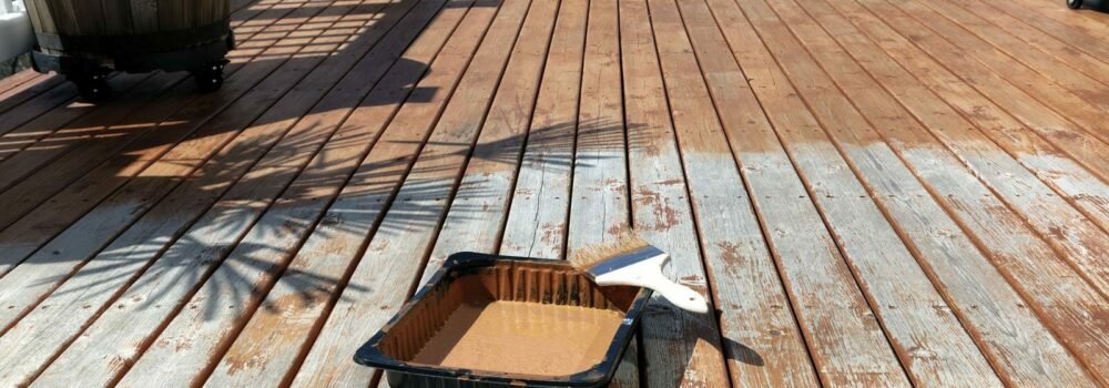 Large paint brush resting on top of tray with stained cedar wood deck boards in background